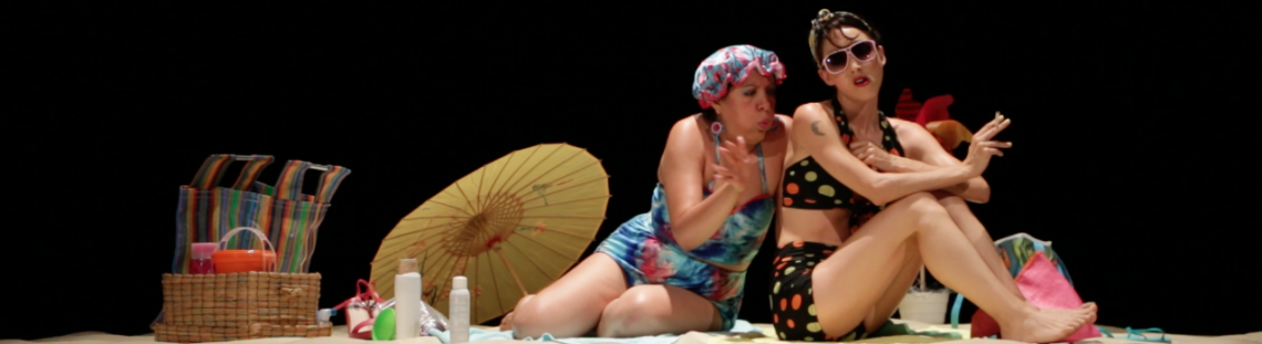 Escenario vestido a manera de playa con dos mujeres en traje de baño conversando.