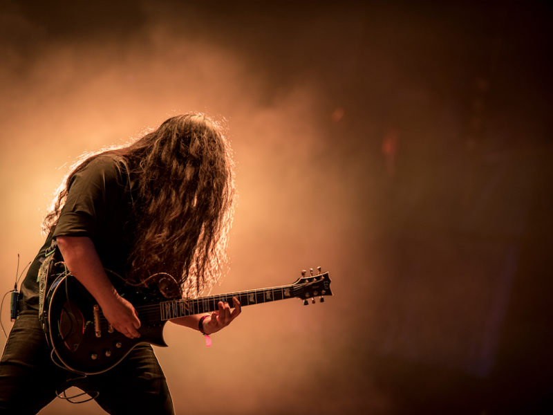 Hombre de pelo largo tocando una guitarra eléctrica con una luz amarilla de fondo, en un evento musical
