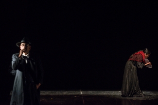 Hombre de negro y sombrero, en la parte posterior del escenario, atras, una mujer se inclina y sostiene papeles en la mano.