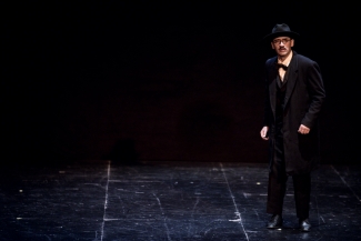 Hombre vestido de negro con gafas y sombrero en escenario oscuro