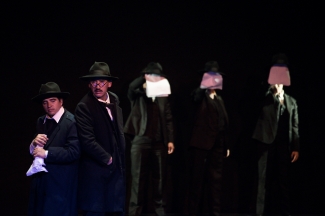 Grupo de hombres vestidos de negro y con sombrero sobre el escenario. Tres de ellos se cubren el rostro con los papeles blancos que sostienen con una mano.