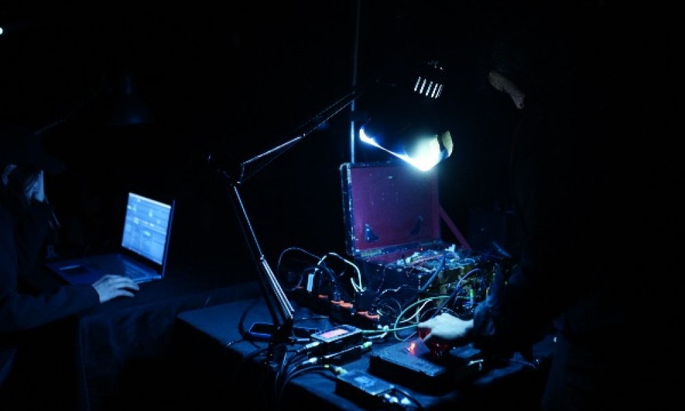 Fotografía en penumbra de una sesión de experimentación sonora. En primer plano, una persona manipula cables y dispositivos electrónicos sobre una mesa, iluminada por una lámpara de escritorio. Al fondo, otra persona trabaja frente a una computadora portátil.