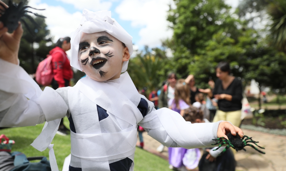 Halloween es protagonista en la agenda del Idartes. Niño disfrazado de momia con dos arañas en las manos juega en el parque de día.