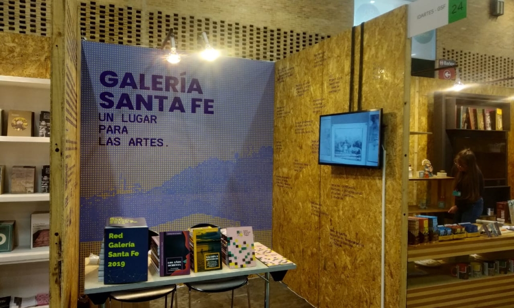 Stand de la Galería Santa Fe en Artbo con libros de arte y una pantalla. Stand de la Galería Santa Fe en Artbo con libros de arte y una pantalla.