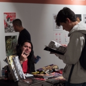 Asistente a las FLA ojea productos de Ave Negra Comics mientras el artista saluda a la camara