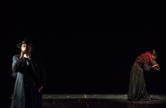 Hombre de negro y sombrero, en la parte posterior del escenario, atras, una mujer se inclina y sostiene papeles en la mano.