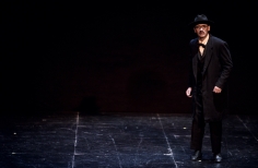 Hombre vestido de negro con gafas y sombrero en escenario oscuro