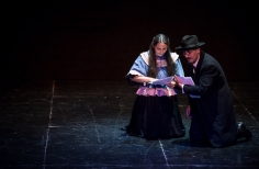 Hombre vesttido de negro formal, con gafas y sombrero arrodillado sobre el escenario con mujer de pelo largo. Anbos leen los papeles que tienen en la mano
