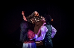 Pareja de actores abrazados. Al fondo una mujer mayor canta o grita.