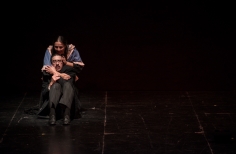 Hombre vestido de negro acurrucado sobre el piso del escenario. Una mujer de pelo largo lo abraza por detras.
