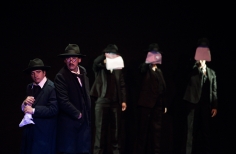 Grupo de hombres vestidos de negro y con sombrero sobre el escenario. Tres de ellos se cubren el rostro con los papeles blancos que sostienen con una mano.