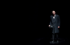 Hombre de negro con sombrero en la mano, con telón oscuro al fondo.