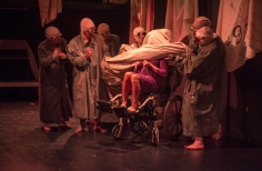 Mujer en silla de ruedas con el rostro cubierto por una manta y hombres con batas alrededor.