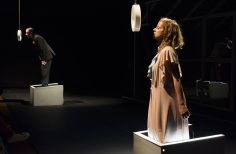 Hombre y mujer actuando, cada uno, con los pies dentro de una especie de caja blanca de madera y frente a un círculo que cuelga del techo.