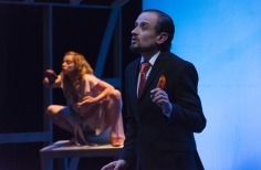 Mujer acurrucada sobre una mesa y hombre actuando.