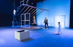 Hombre y mujer actuando en escenario con equeleto de casa, impermeable y zapatos de invierno.