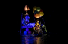 Mujer en cama flotante con globos y hombre con bicicleta sobre el lago.