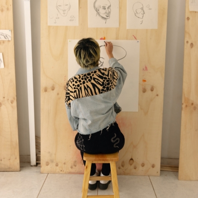 Fotografía de persona sentada en una butaca dibujando a mano alzada sobre papel colgado en pared.