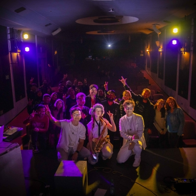 Artistas Shimura Cinema posando con público de fondo después de su concierto.