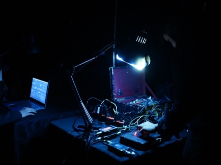 Fotografía en penumbra de una sesión de experimentación sonora. En primer plano, una persona manipula cables y dispositivos electrónicos sobre una mesa, iluminada por una lámpara de escritorio. Al fondo, otra persona trabaja frente a una computadora portátil.
