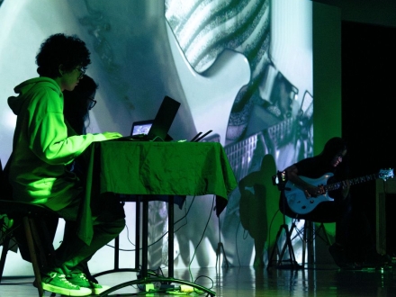 Un escenario oscuro con una pantalla donde se proyectan imágenes en sepia. En el escenario están tres personas. Dos de ellos son músicos. La tercera es una Vj. Los tres realizan una improvisación sonora y visual en vivo