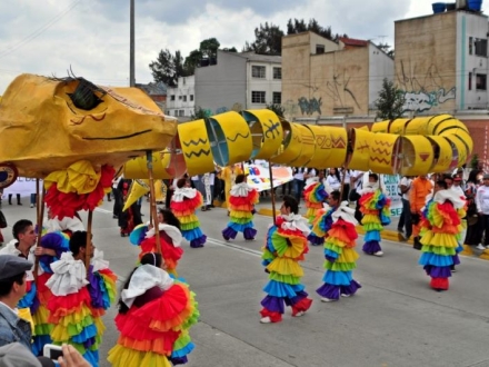 Comparsa en espacio público, jóvenes danzando con vestidos y una maqueta sobre sus cabezas.