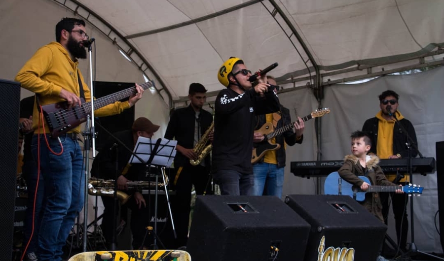 Agrupación musical realizando una presentación en vivo sobre una tarima en espacio abierto, en la cual se ven siete hombres adultos, y un niño sosteniendo una guitarra azul.