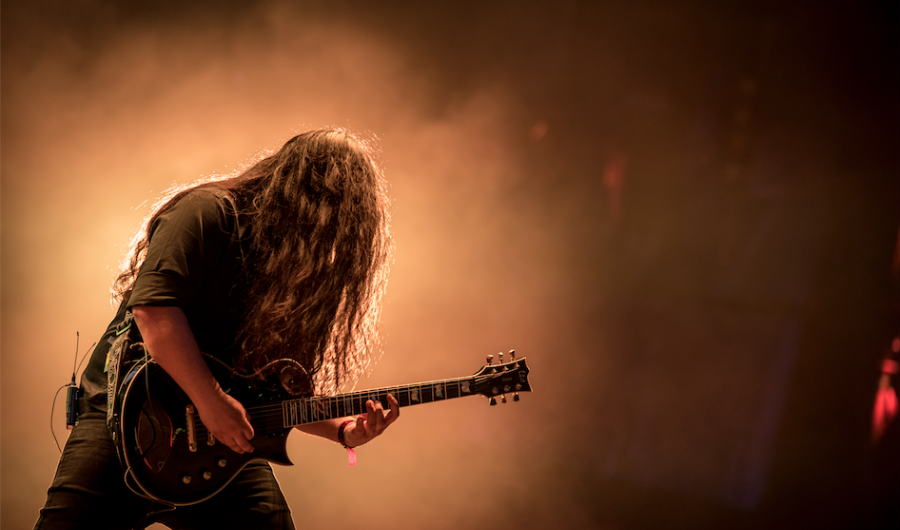 Hombre de pelo largo tocando una guitarra eléctrica con una luz amarilla de fondo, en un evento musical