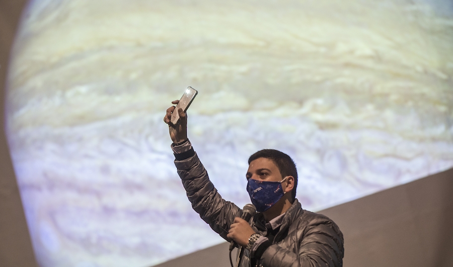 Hombre levantando la mano con un celular con una proyección de Júpiter de fondo.
