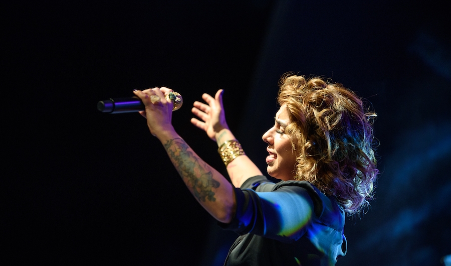 Una mujer cantante alza las manos hacia el público en fondo negro. 