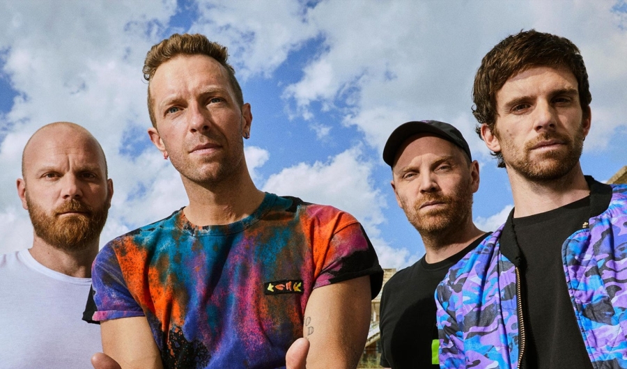 Integrantes de Coldplay con cielo azul y nubes de fondo.