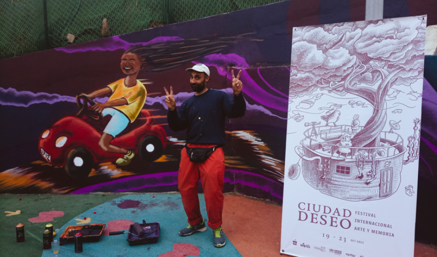 Artista posando con su graffiti detrás y el afiche del Festival Ciudad Deseo de día.