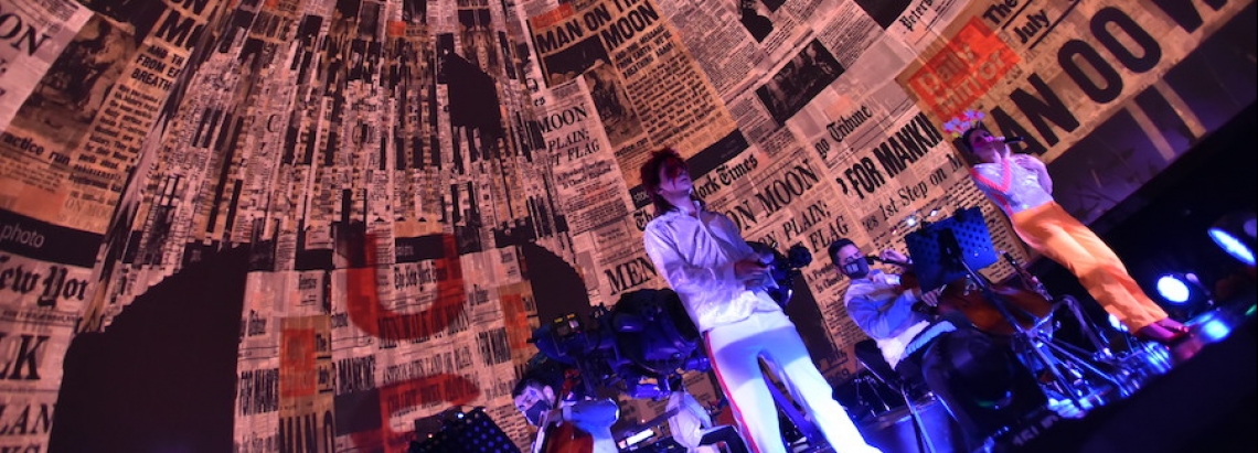 Hombre vestido como David Bowie en la tarima del domo con proyecciones de periódicos en el fondo. 