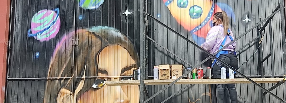 una mujer sobre un andamio pinta la fachada de un Centro de Formación. El grafiti consiste en un rostro de mujer y una nave espacial.