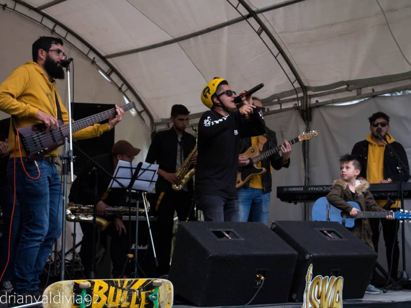 Agrupación musical realizando una presentación en vivo sobre una tarima en espacio abierto, en la cual se ven siete hombres adultos, y un niño sosteniendo una guitarra azul.