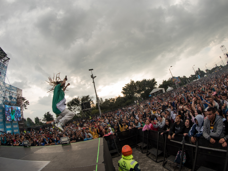 Hombre de pelo largo saltando en la tarima de un festival de música ante un público masivo.