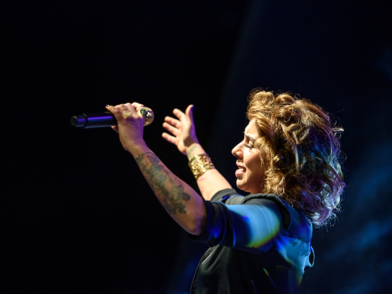 Una mujer cantante alza las manos hacia el público en fondo negro. 