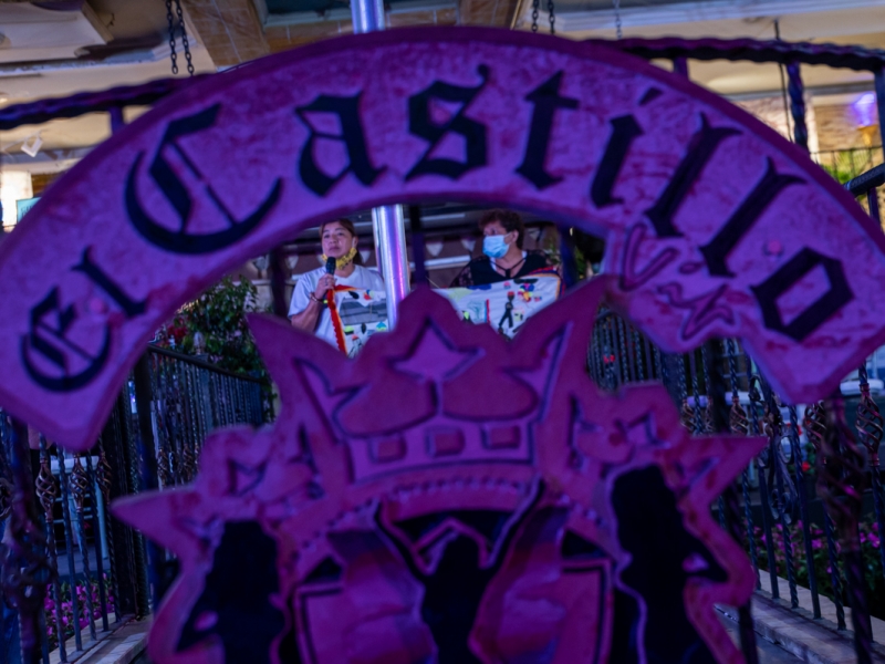 Letrero del castillo en primer plano, al fondo dos señoras con bordados, de noche.