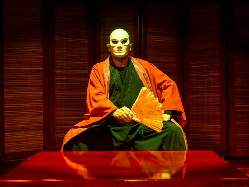 Hombre con una máscara y un kimono sentado en la tarima del escenario 