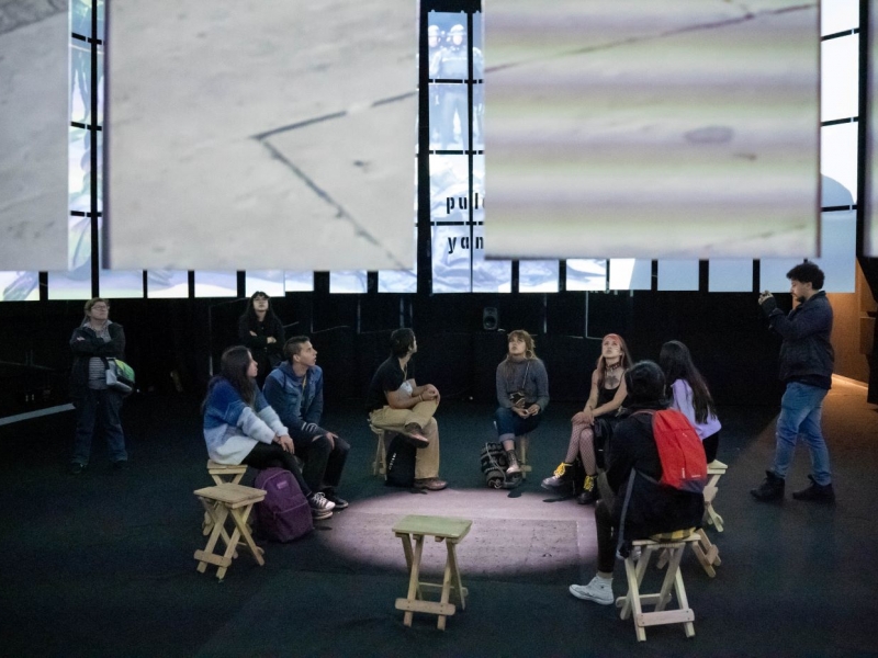 Varias personas sentadas en círculo  viendo una presentación de mapping en una sala oscura