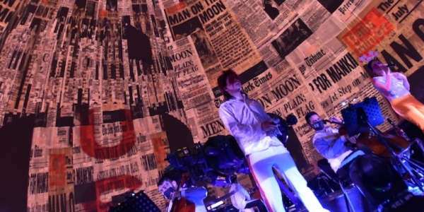 Hombre vestido como David Bowie en la tarima del domo del Planetario con imágenes de periódicos en el fondo. 
