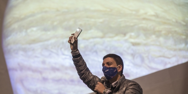 Hombre levantando la mano con un celular con una proyección de Júpiter de fondo. 