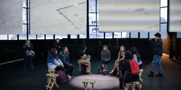 Varias personas sentadas en círculo  viendo una presentación de mapping en una sala oscura