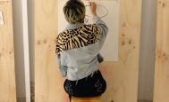 Fotografía de persona sentada en una butaca dibujando a mano alzada sobre papel colgado en pared.
