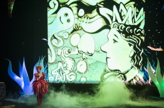 Actriz de rojo actuando sobre un escenario con humo verde e imagen de dibujos como fondo