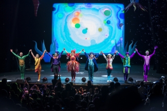 Actores recibiendo aplausos sobre el escenario con fondo de burbujas coloridas