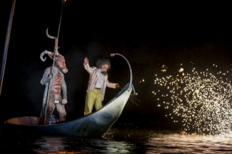 Actores sobre góndola azul sobre lago iluminado.