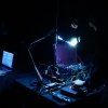 Fotografía en penumbra de una sesión de experimentación sonora. En primer plano, una persona manipula cables y dispositivos electrónicos sobre una mesa, iluminada por una lámpara de escritorio. Al fondo, otra persona trabaja frente a una computadora portátil.