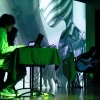 Un escenario oscuro con una pantalla donde se proyectan imágenes en sepia. En el escenario están tres personas. Dos de ellos son músicos. La tercera es una Vj. Los tres realizan una improvisación sonora y visual en vivo